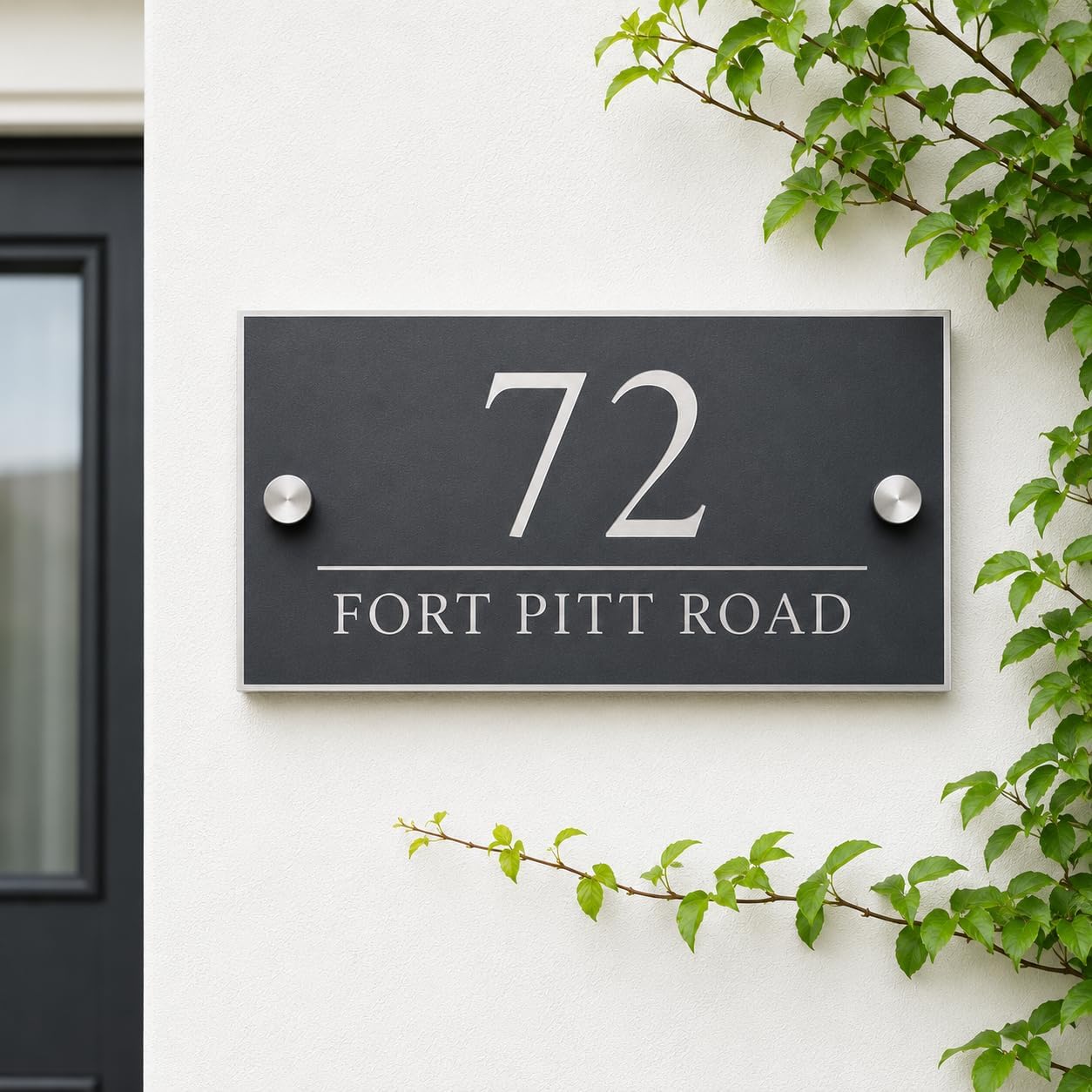 A modern black house number plaque with silver lettering reading '72 FORT PITT ROAD' mounted on a white exterior wall, with green vines framing the scene.