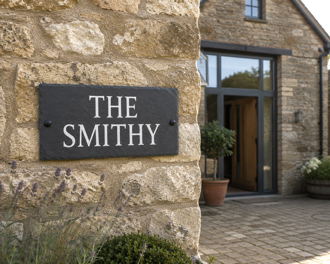 A dark slate sign reading 'THE SMITHY' in white lettering, mounted on a rustic stone wall beside a stone building with a glass door and potted plants.