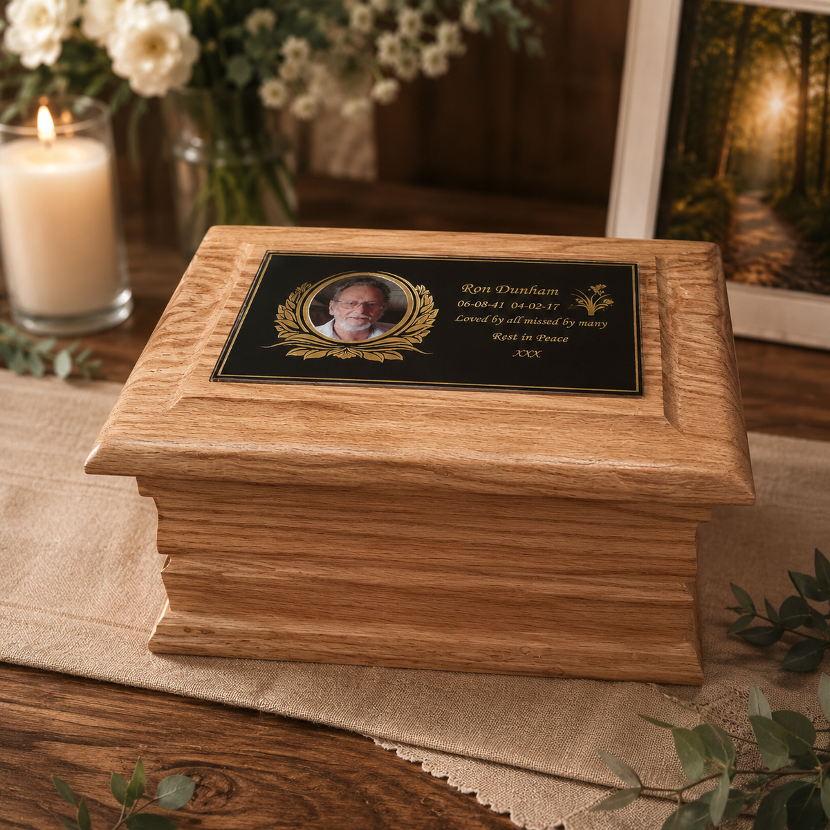 A wooden keepsake box or urn with a black memorial plaque featuring a portrait photo and gold text, displayed on a tabletop with a candle, flowers, and a framed photo in the background.