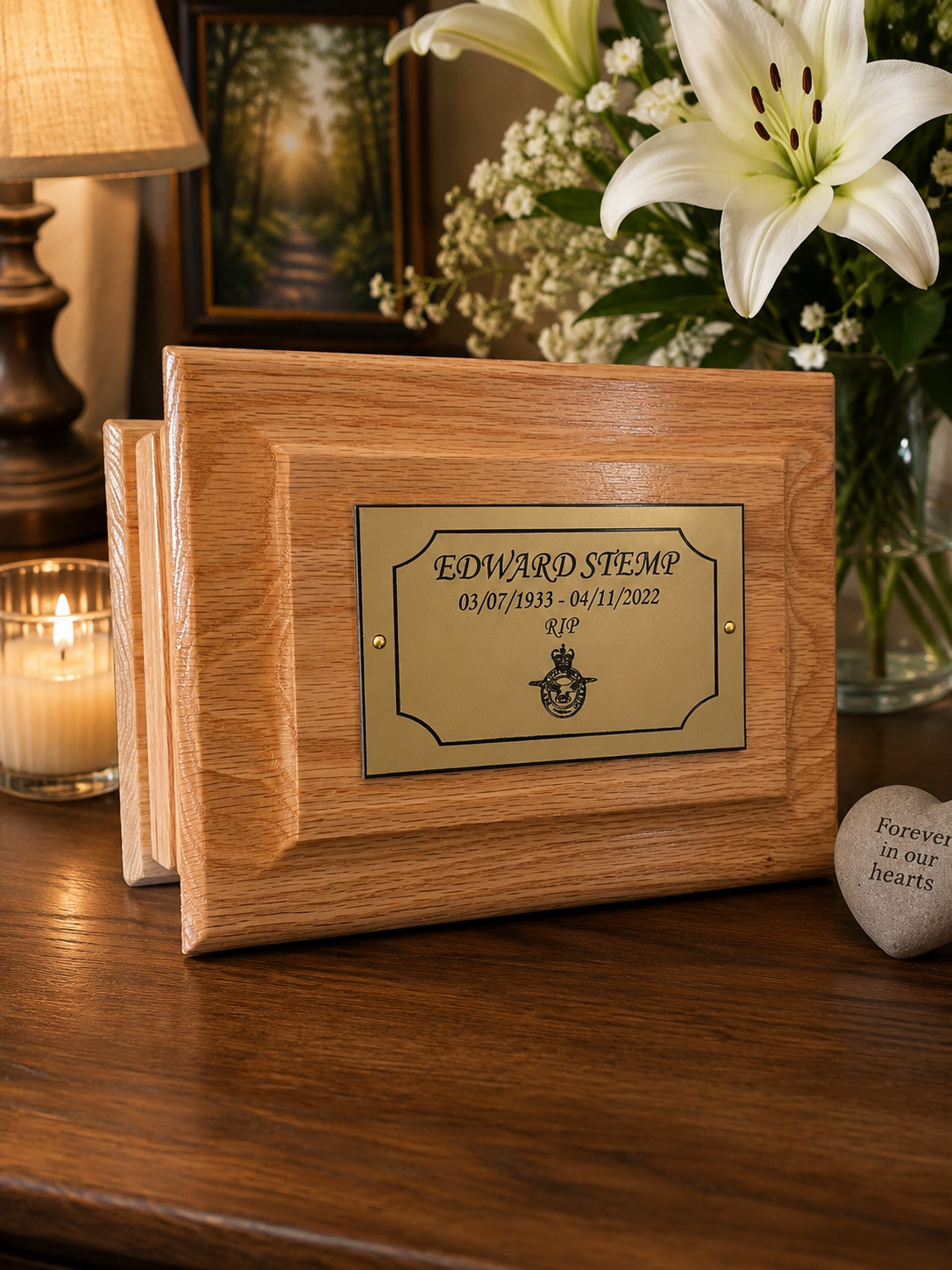 A wooden keepsake urn or memorial box with a gold-toned engraved plaque reading 'EDWARD SIEMP' and dates, displayed on a wooden table with flowers, a lamp, and a candle in the background.