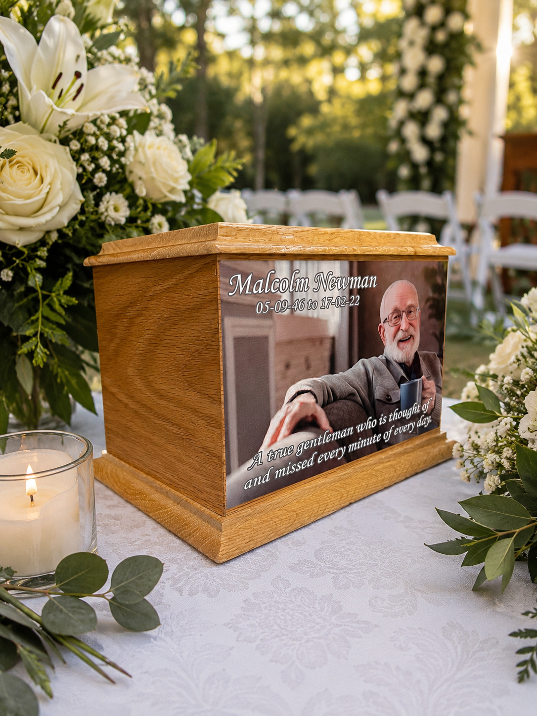 A wooden cremation urn with a custom photo print of a man named Malakin Newman, displayed on a white tablecloth surrounded by white flowers and a lit candle.