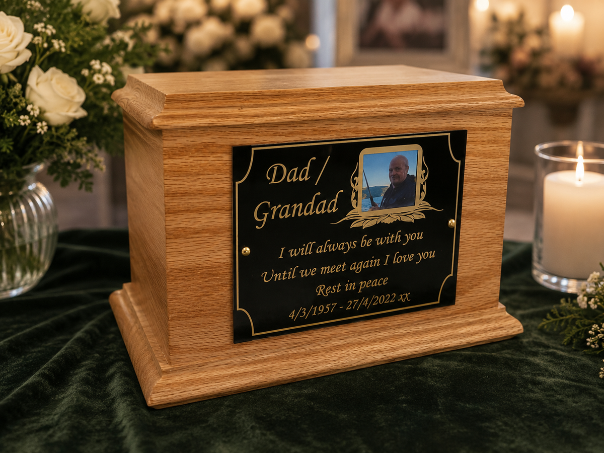 A wooden keepsake urn with a black engraved plaque reading 'Dad / Grandad' and a photo of a man, displayed on a dark green velvet cloth with flowers and a lit candle in the background.