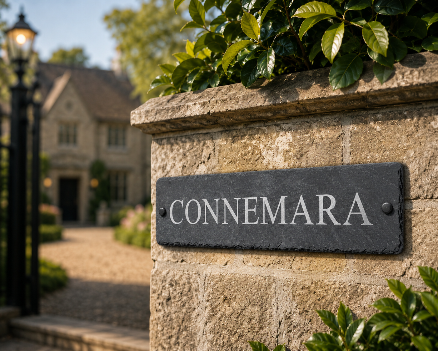 A rectangular dark slate sign with the name 'CONNEMARA' in white capital letters, mounted on a light beige stone wall with green foliage above and below, with a blurred house and garden entrance in the background.