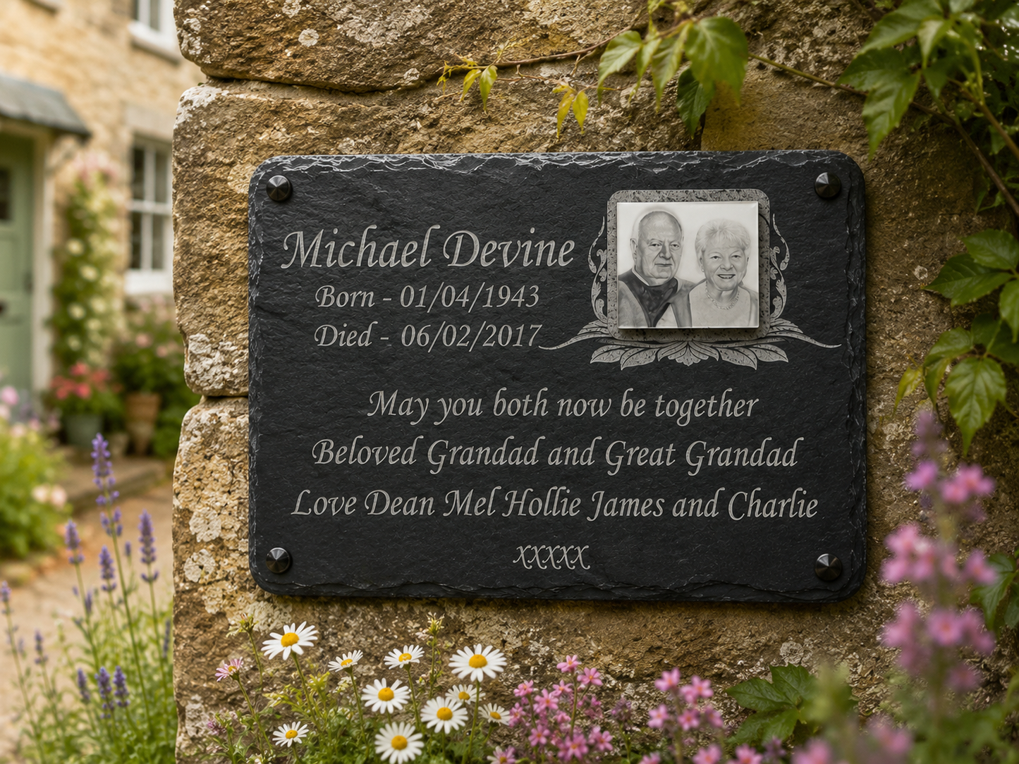 A personalized black slate memorial plaque mounted on a rustic stone wall, featuring engraved white text and a small portrait photo of a grandfather and grandmother. The plaque is surrounded by green ivy and small white and pink flowers, with a blurred stone building in the background.