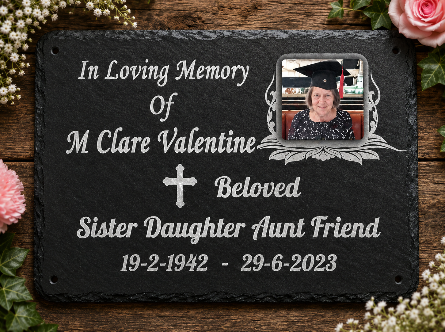 A personalized black slate memorial plaque with white engraved text reading 'In Loving Memory of M Clare Valentine, Beloved Sister Daughter Aunt Friend 19-2-1942 - 29-6-2023,' plus a photo frame showing a woman in a graduation cap. The plaque is styled on a rustic wooden surface with pink flowers and green leaves around the edges.