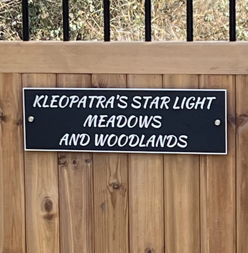 Aluminium sign with text 'KLEOPATRA'S STAR LIGHT MEADOWS AND WOODLANDS' on a wooden fence.