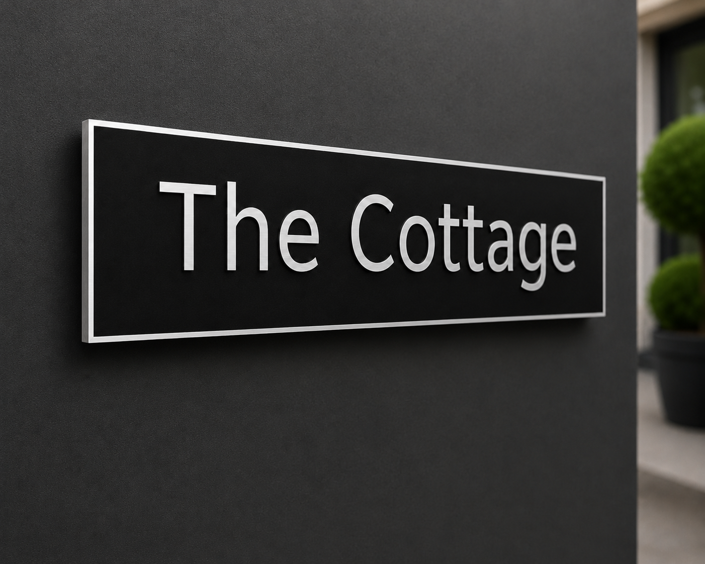 A rectangular black sign with a thin white border and the words 'The Cottage' in white serif lettering, mounted on a dark gray exterior wall with a blurred building and greenery in the background.