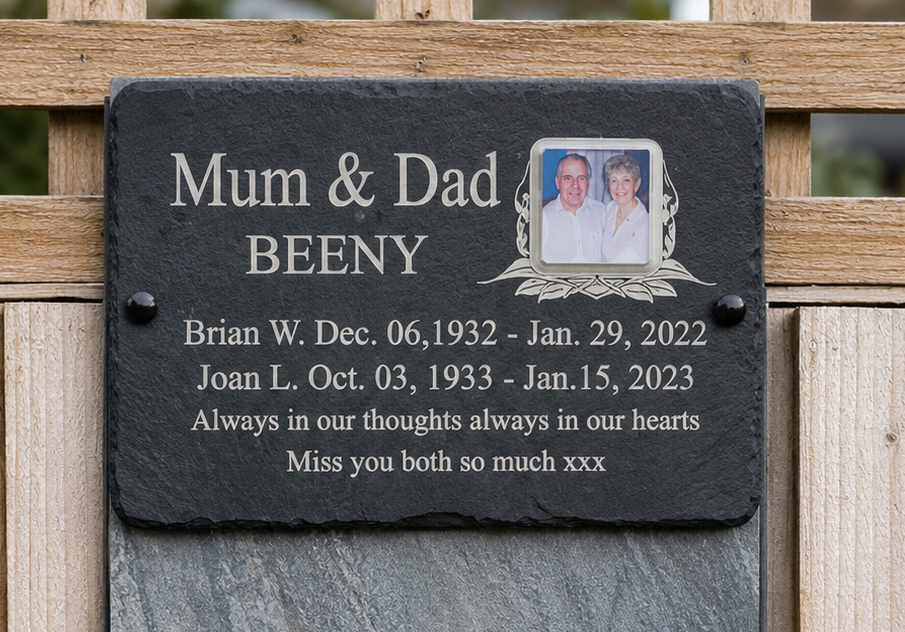 A personalized black slate memorial plaque mounted on a wooden fence, featuring engraved white text for 'Mum & Dad BEENY' with birth and death dates and remembrance words, plus a small inset color photo of a couple.