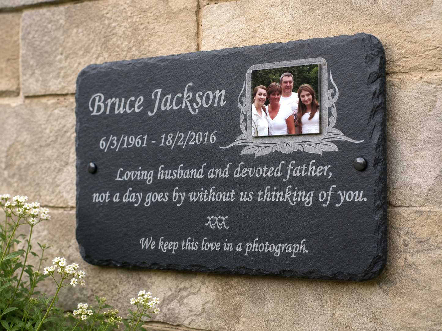 Personalized black slate memorial plaque mounted on a beige stone wall, engraved with the name Bruce Jackson, birth and death dates, a family photo, and remembrance text.