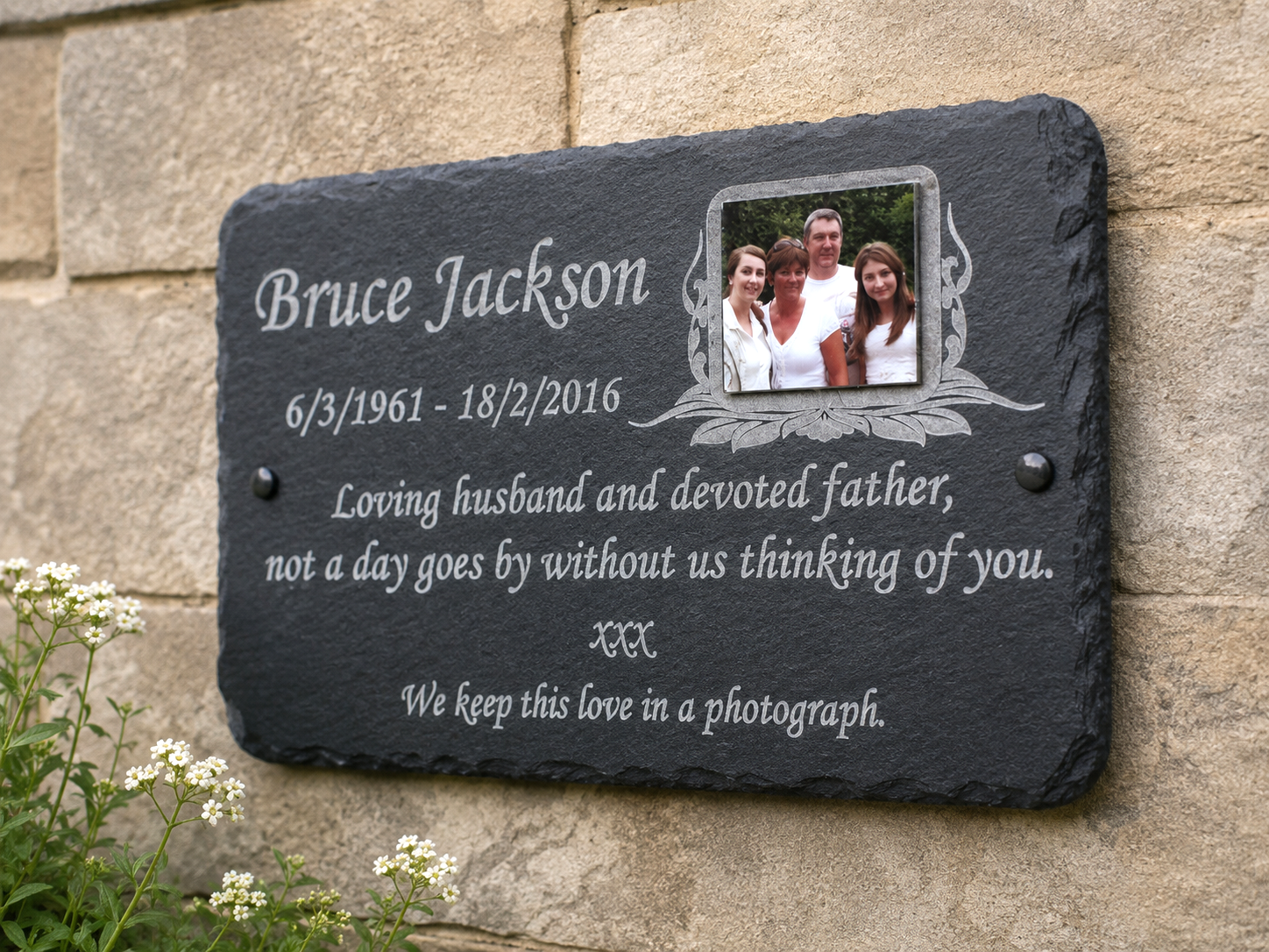 Personalized black slate memorial plaque mounted on a beige stone wall, engraved with the name Bruce Jackson, birth and death dates, a family photo, and remembrance text.