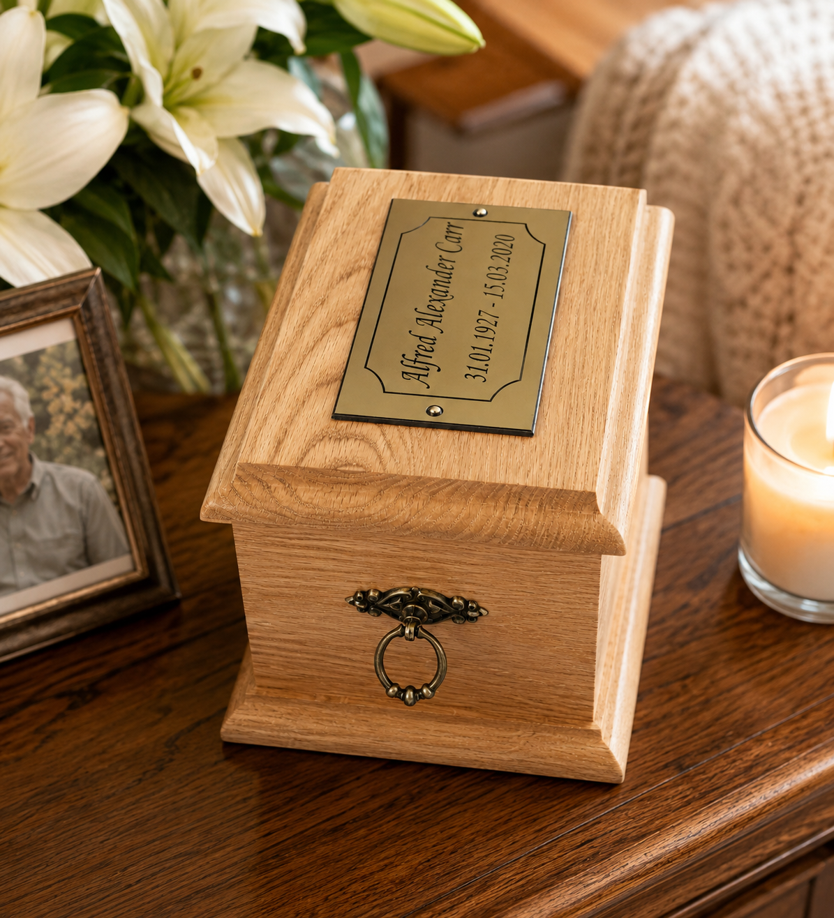 A wooden keepsake urn box with a brass nameplate and decorative metal ring pull, displayed on a wooden table with a lit candle, flowers, and a framed photo in a home setting.