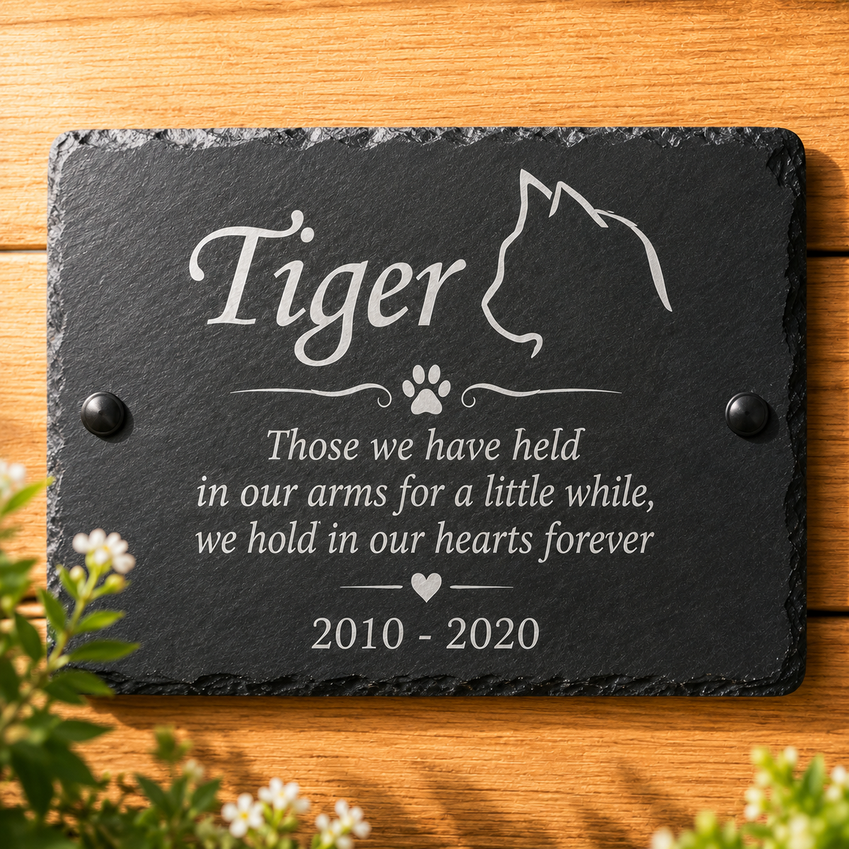 Personalized black slate pet memorial plaque engraved with the name 'Tiger', a cat silhouette, paw print, and remembrance text, displayed on a wooden surface with small white flowers in the foreground.