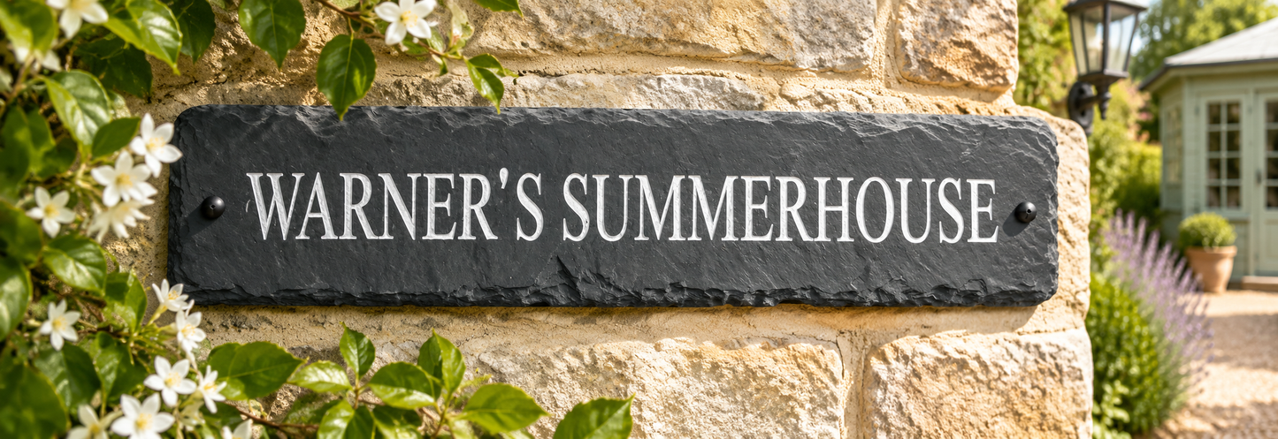 A rectangular black slate house sign with white lettering that reads 'WARNER'S SUMMERHOUSE,' mounted on a light tan stone wall with green foliage and small white flowers around it.