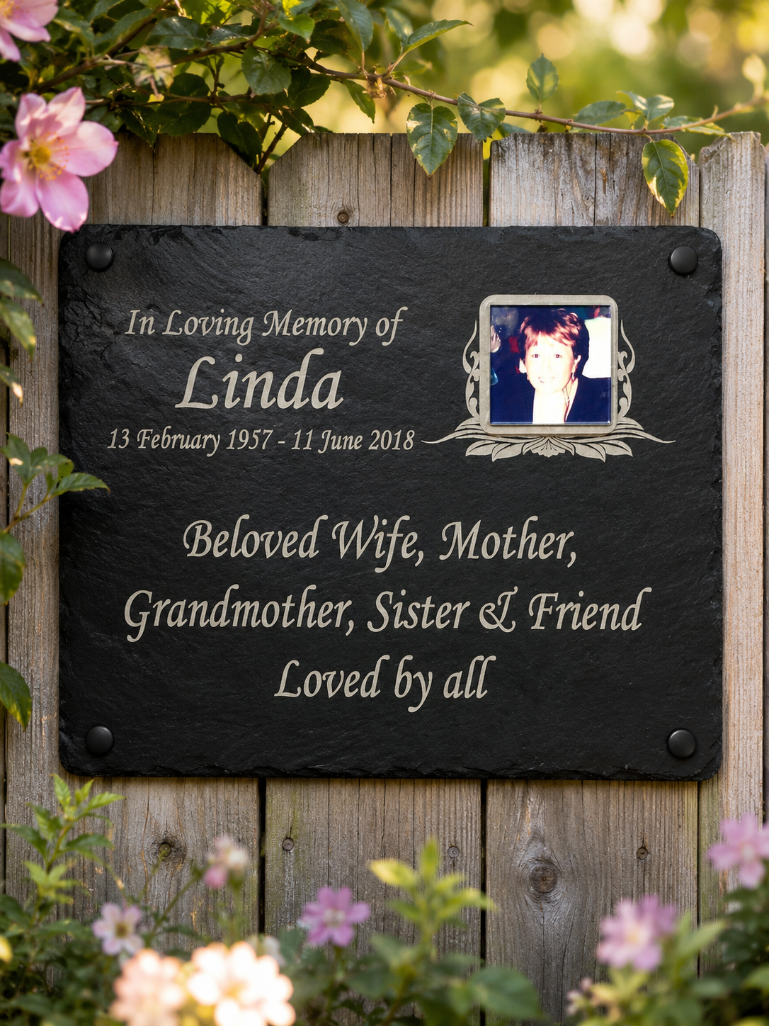 A personalized black slate memorial plaque mounted on a weathered wooden fence, surrounded by green leaves and pink flowers. The plaque features engraved white text reading 'In Loving Memory of Linda' with dates and 'Beloved Wife, Mother, Grandmother, Sister & Friend Loved by all', alongside a small portrait photo of Linda.