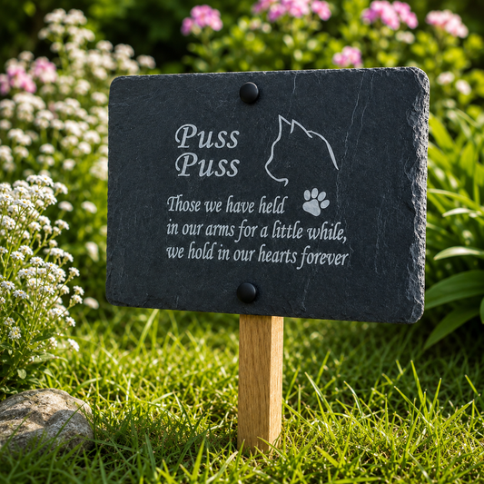 A rectangular dark slate pet memorial garden sign with engraved white text reading 'Puss Puss' and a paw print, mounted on a wooden stake in a grassy garden with flowers in the background.