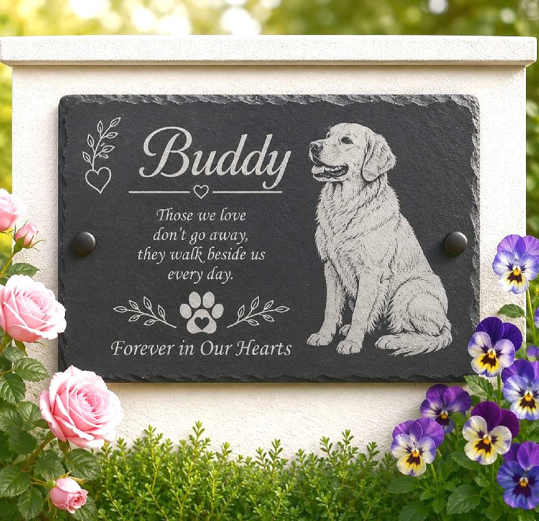 Personalized black slate pet memorial plaque engraved with a golden retriever illustration and the name 'Buddy,' mounted on a cream-colored stone wall with pink roses and purple flowers in the foreground.