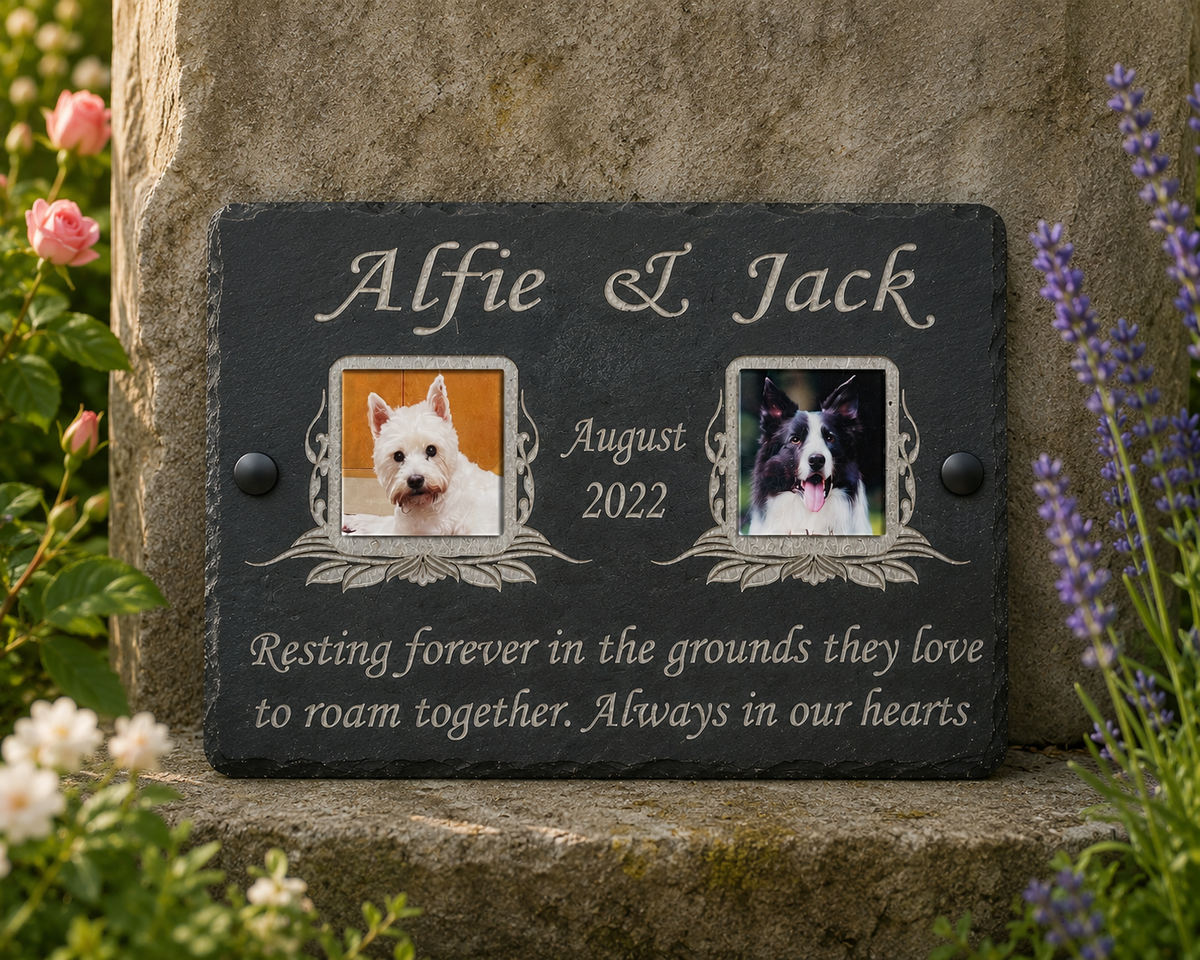Personalized black slate pet memorial plaque with engraved silver lettering, two dog photo frames, and a remembrance quote, mounted on a stone wall with flowers around it.