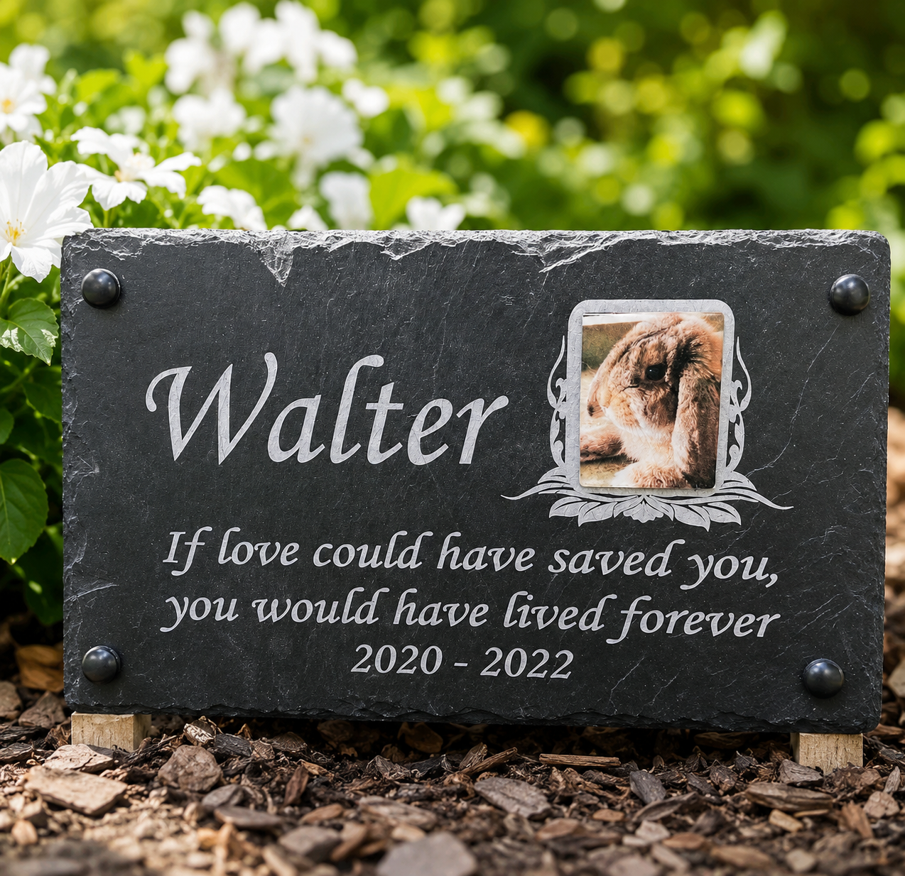 Personalized black slate garden memorial plaque with engraved white text reading 'Walter' and a photo of a cat, placed outdoors on mulch with green foliage and white flowers in the background.