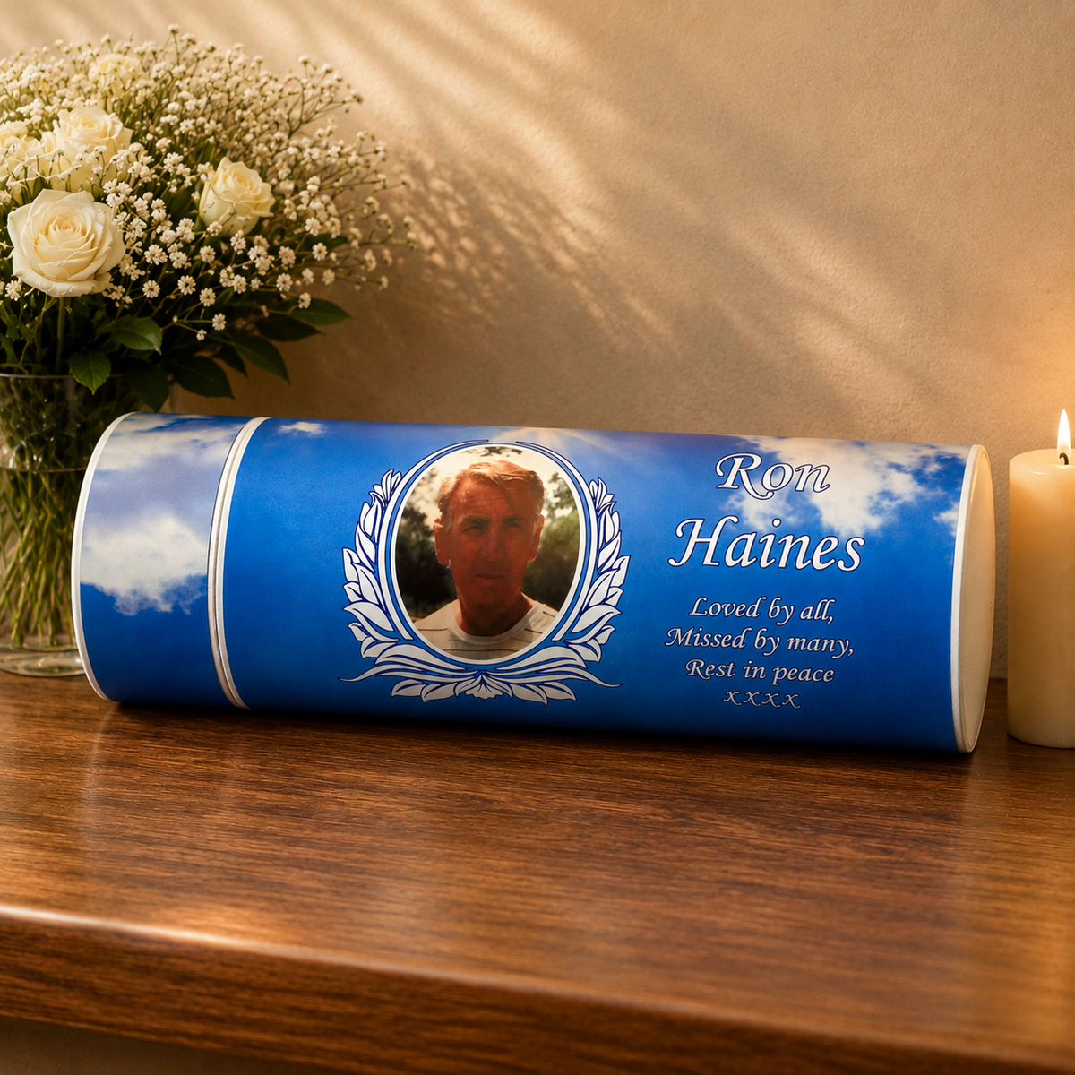 A personalized blue cylindrical urn candle with a photo of a man, the name 'Rom Haines', and a quote, displayed on a wooden table next to a bouquet of white roses and a lit candle.