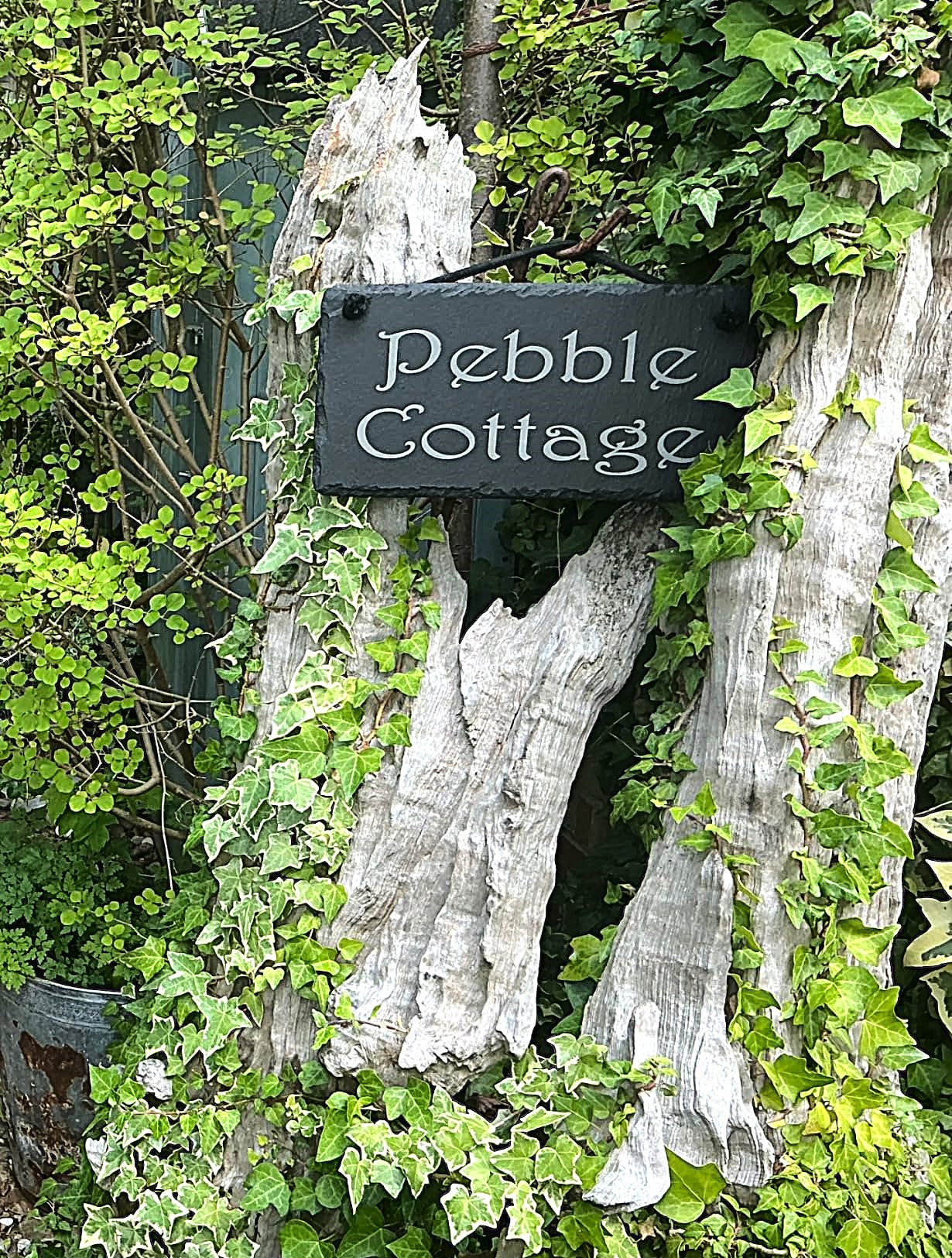 A rustic black wooden sign reading 'Pebble Cottage' in white script, mounted on weathered tree trunks surrounded by green ivy and foliage outdoors.