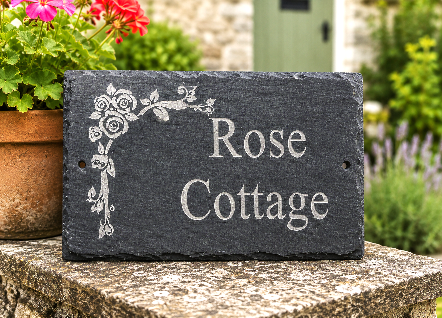 A rectangular dark slate house sign with engraved white floral corner decoration and the words 'Rose Cottage,' displayed outdoors on a stone surface with flowers and greenery in the background.