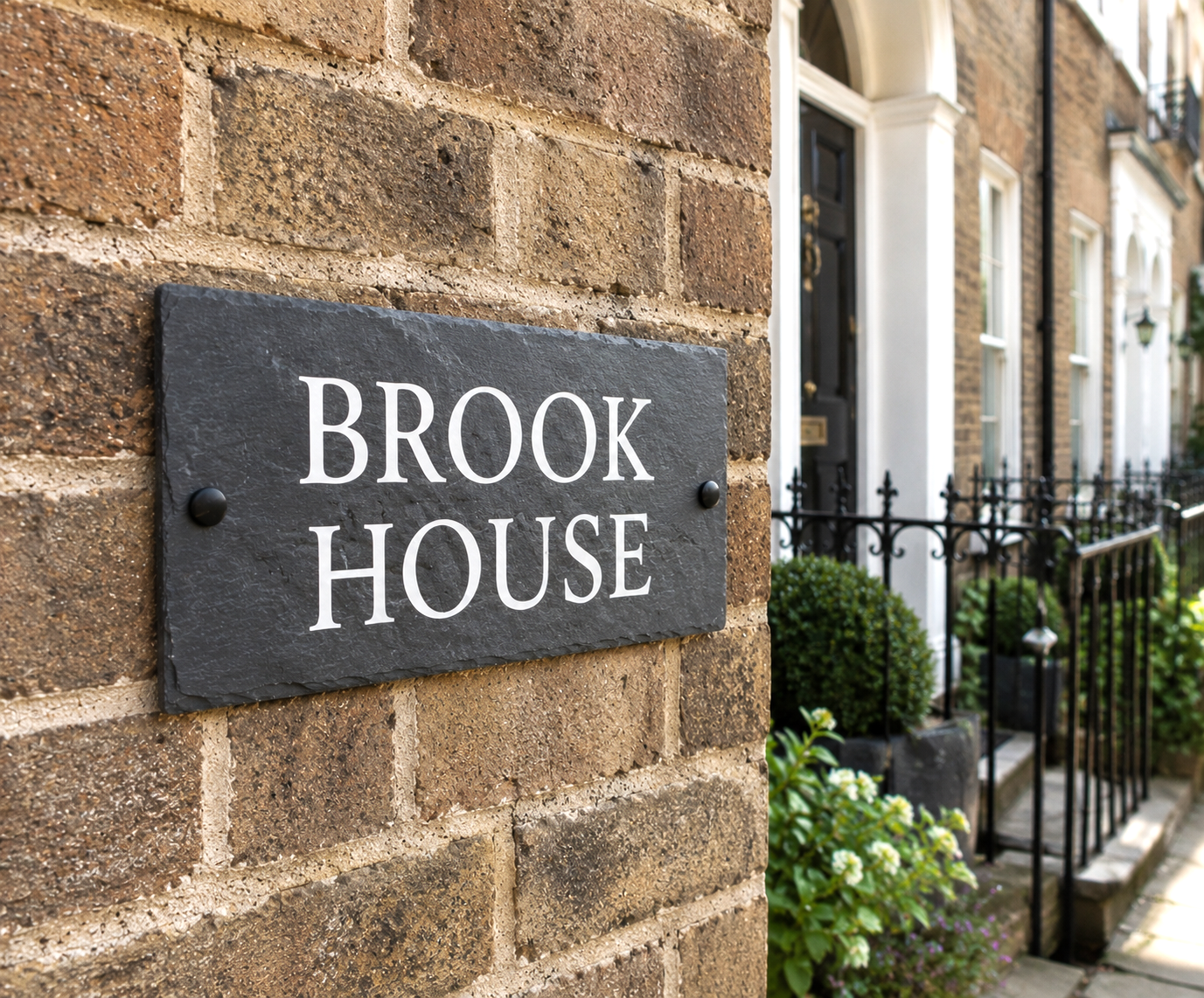 A rectangular dark slate house sign with white serif text reading 'BROOK HOUSE' mounted on a brown brick wall, with a row of similar houses and a black iron fence visible in the background.
