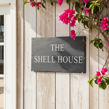 Slate sign on a wooden wall with pink flowers above it, labeled 'The Shell House'.