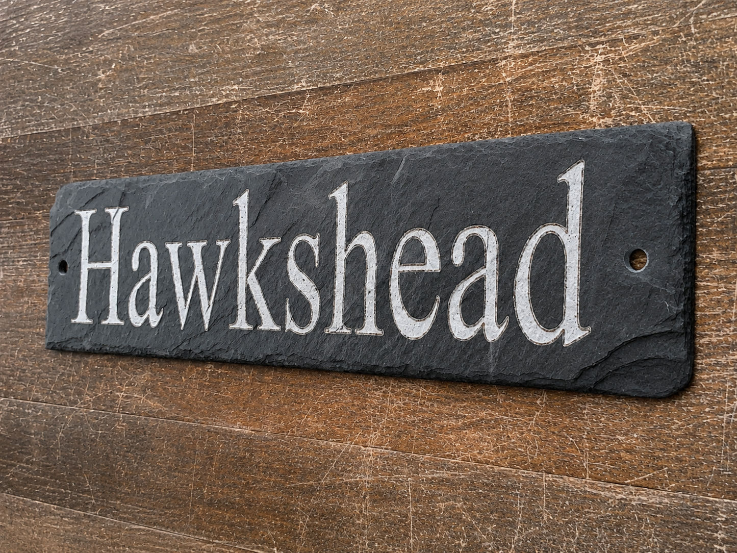 A rectangular black slate sign with the word 'Hawkshead' in white serif lettering, mounted on a rustic brown wooden surface.