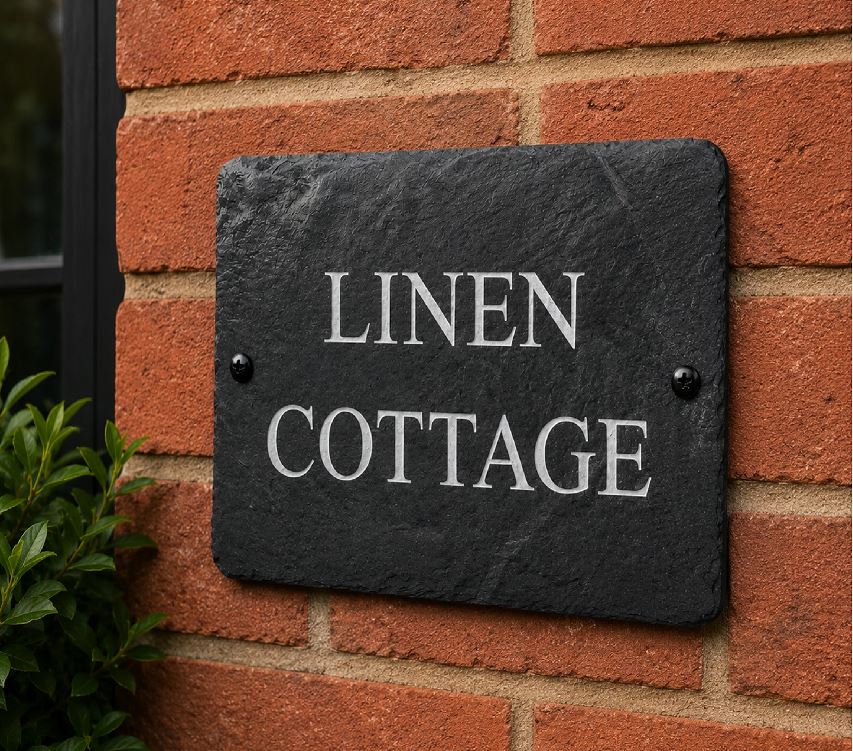 A rectangular black slate house sign with the words 'LINEN COTTAGE' in white serif lettering, mounted on a red-orange brick wall with two small black screws.
