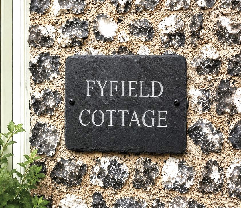 A rectangular black slate house plaque with the words 'FYFIELD COTTAGE' in raised silver lettering, mounted on a rough stone wall.