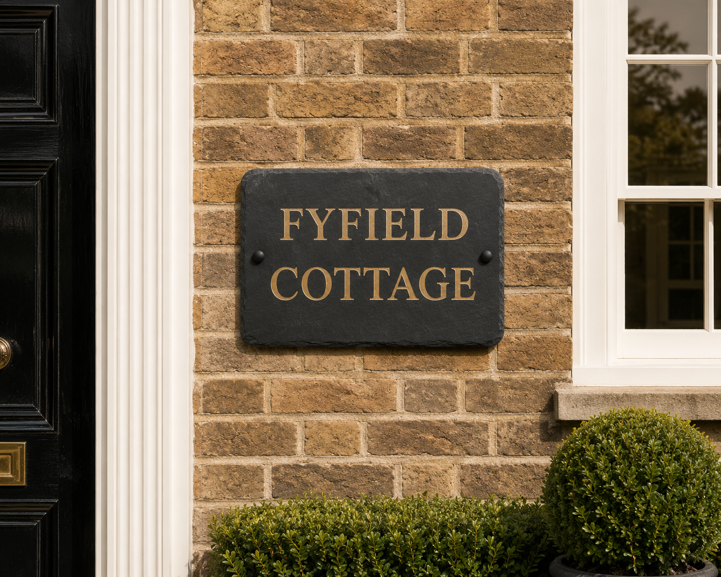 A rectangular black slate house plaque with gold lettering reading 'FYFIELD COTTAGE' mounted on a tan brick wall next to a white-framed window.