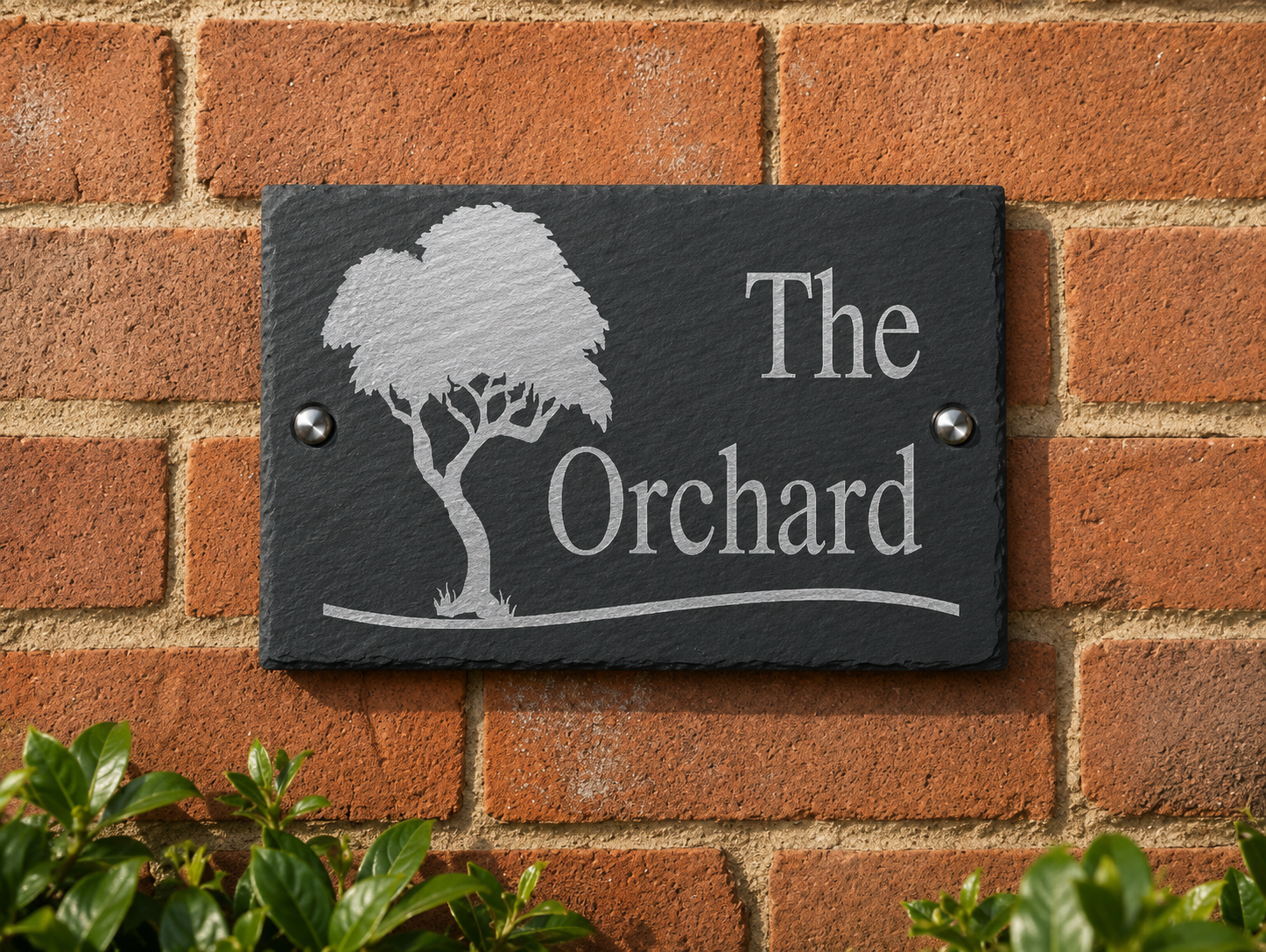 A rectangular dark slate plaque mounted on a red brick wall, featuring a white engraved tree silhouette and the words 'The Orchard' with a curved line beneath. Small metal screws secure the plaque at two sides.