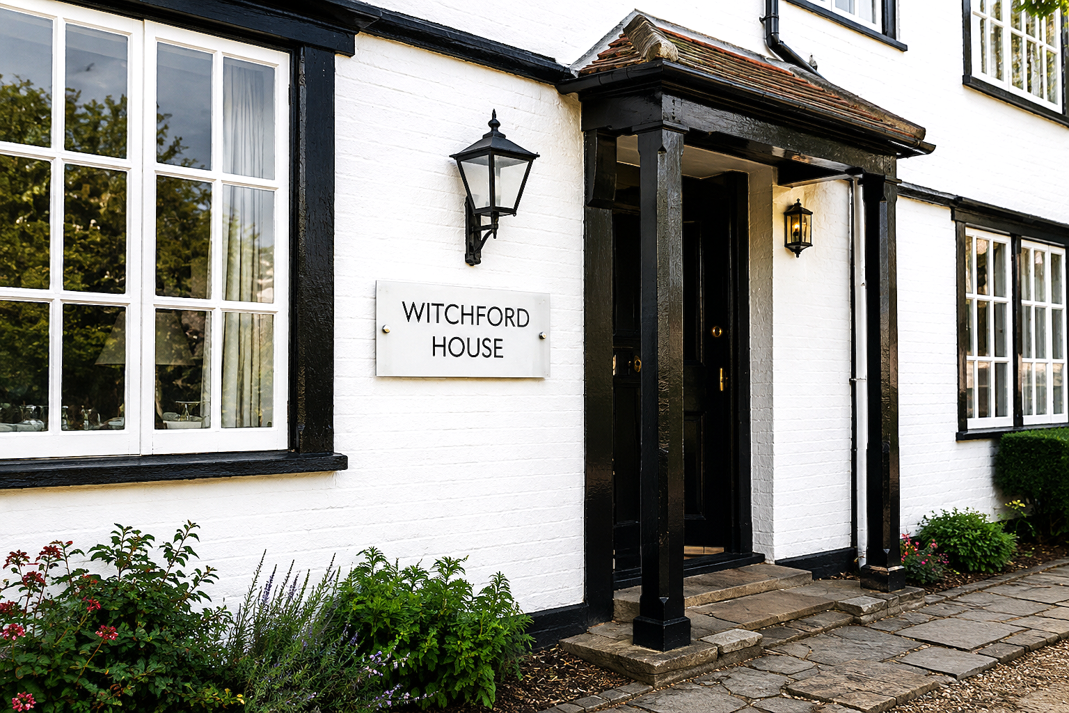 White brick house exterior with black trim, a central entrance porch with dark posts, wall-mounted lanterns, and a sign reading 'WITCHFORD HOUSE'.