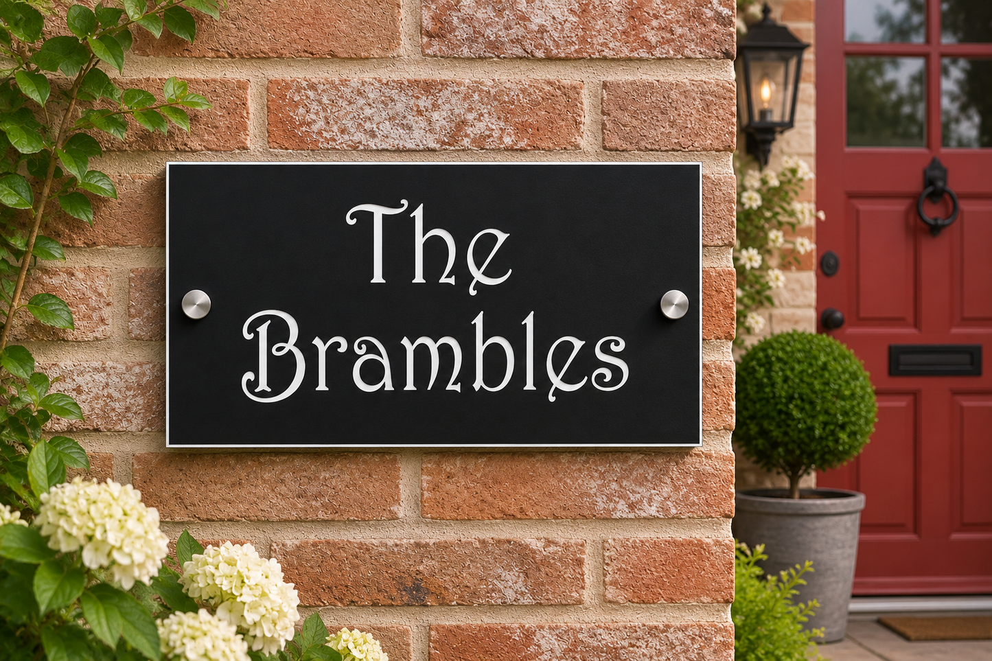 A rectangular black house sign with silver metal mounting hardware, featuring the white script text 'The Brambles' mounted on a red brick wall next to a red front door.