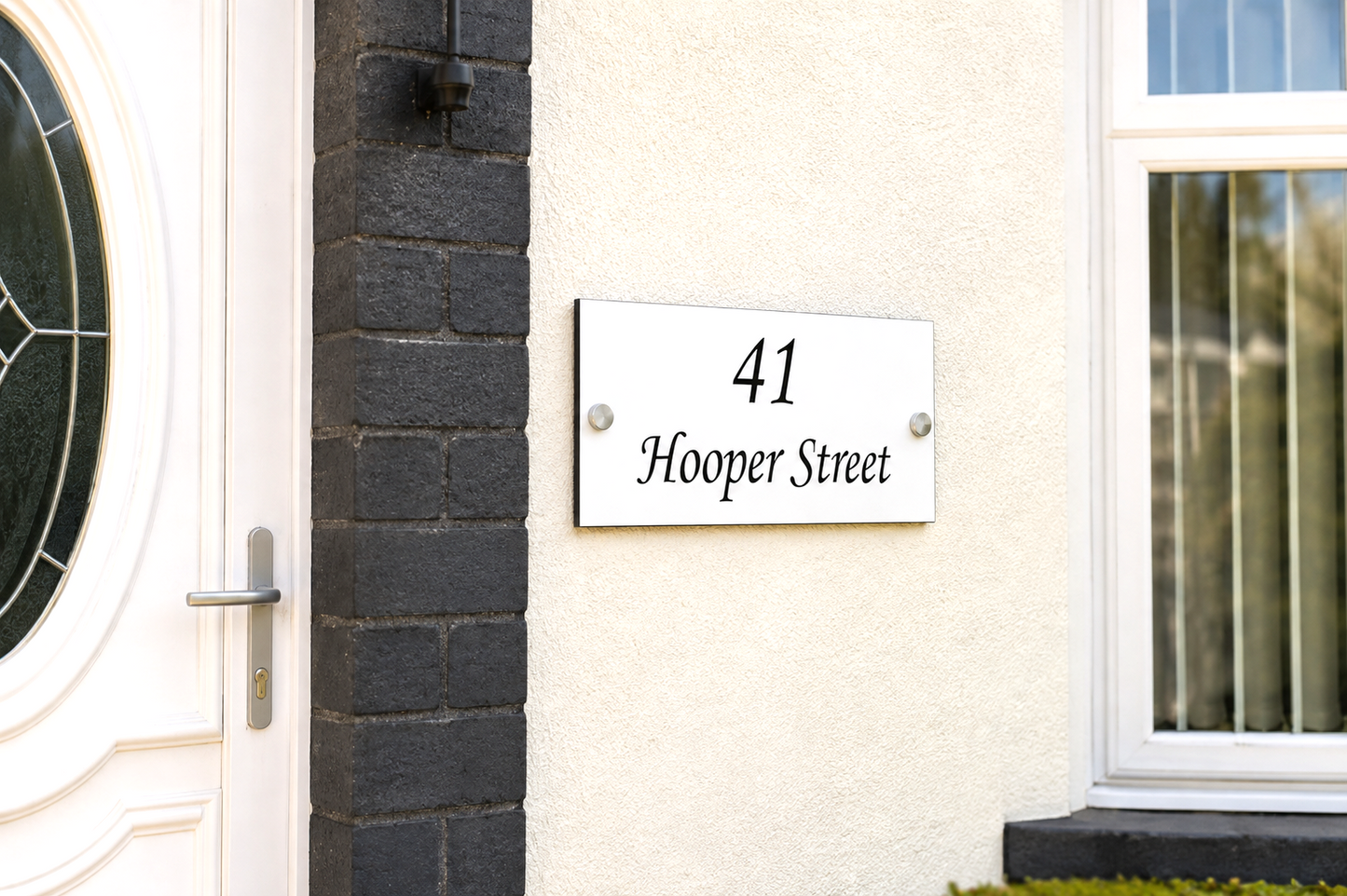 A white rectangular house number plaque reading '41 Hooper Street' mounted on a cream-colored stucco wall next to a dark gray brick pillar and a white front door.