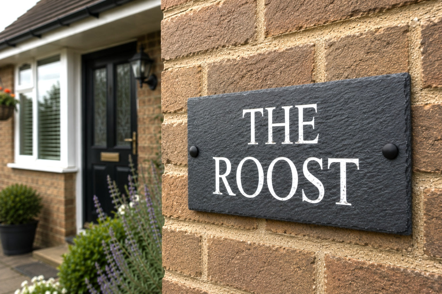 A rectangular black slate house sign with white serif text reading 'THE ROOST' mounted on a tan brick wall beside a house entrance.
