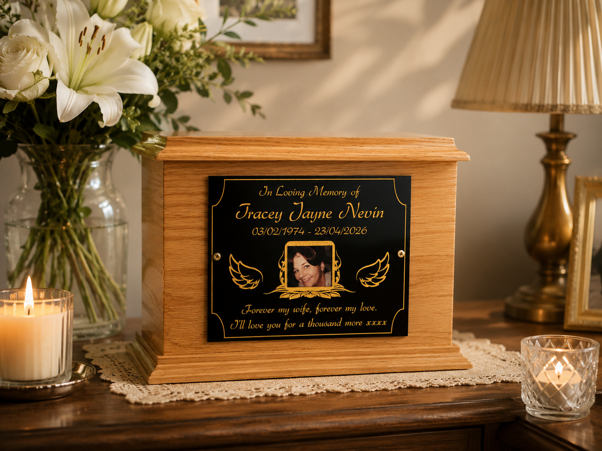 A wooden cremation urn with a black memorial plaque featuring gold lettering and a photo frame, displayed on a wooden table with a lit candle, white lilies, and a table lamp in a warm indoor setting.