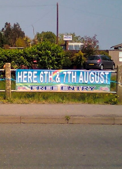 A promotional banner for an event, placed on a fence, with the text 'HERE 6th & 7th AUGUST FREE ENTRY' printed on it.